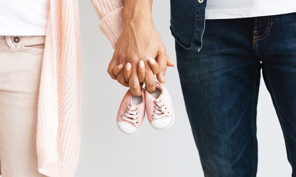 Closeup of pregnant couple carrying small baby shoes while holding hands together, unrecognizable loving man and woman expecting baby, enjoying future parenthood, cropped image, free space